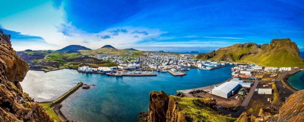 Panoramic view of the Westman Islands in Iceland, featuring a coastal town with colorful buildings, a bustling harbor, rugged cliffs, and clear blue waters under a sunny sky.